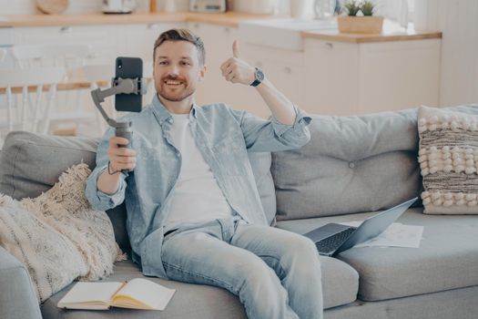 Confident young male freelancer showing thumbs up gesture to his client to inform him that everything is going well through phone video call, sitting on couch holding gimbal with phone
