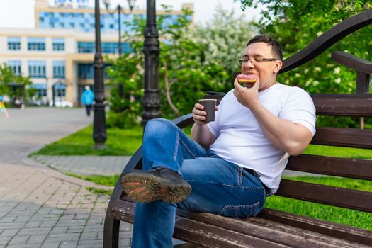 A man in a white t-shirt and jeans is enjoying a delicious donut with a drink while sitting on a park bench in summer