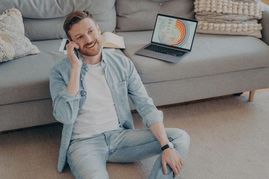 Young man working remotely from home calling project partner to tell him he finished his part of work, gathered enough data, open notebook on couch showing information graphs