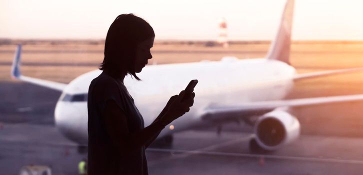 A young girl is texting on a smartphone before boarding a plane with friends, a traveler goes on a trip, a silhouette on the background of the airplane at sunset.