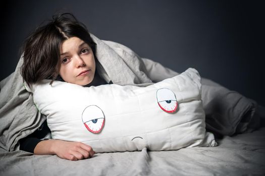 Girl hugs a pillow with tired red eyes and a sad expression on her face, hides under the covers. Woman is worried about insomnia, poor sleep, stress, in her hands a creative pillow with paper eyes.