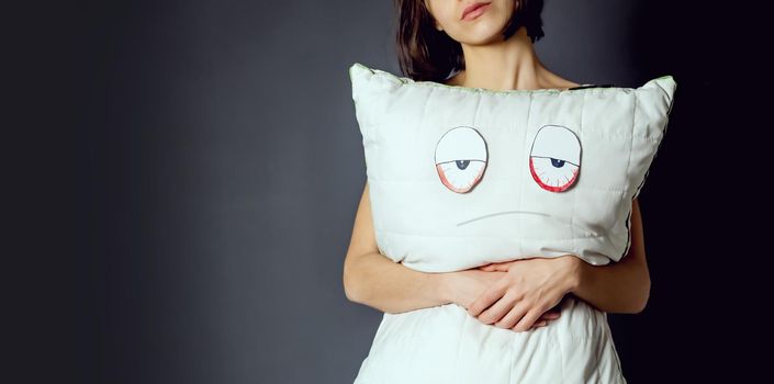 Young girl holds a pillow, on which a displeased face and tired eyes are drawn. Creative idea about sleep problems, insomnia and the importance of quality orthopedic materials.
