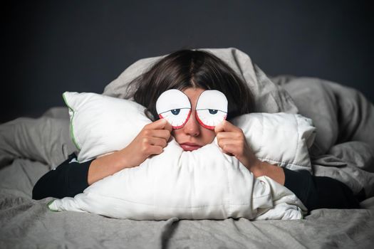 A funny girl hugs a pillow, holds drawn creative tired red eyes in her hands, covered with a blanket. A woman is worried about insomnia, poor sleep, stress.