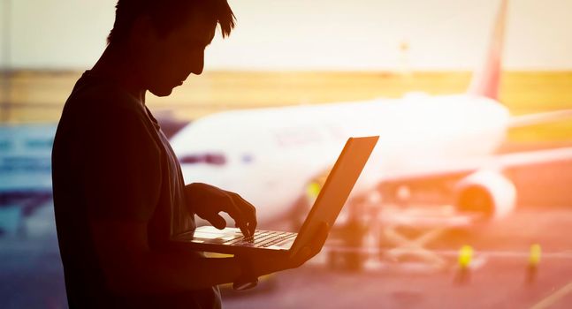 A young man is working on a laptop at the airport while waiting to board the plane. A man is engaged in business, buys tickets, studies and communicates via the Internet at sunset.