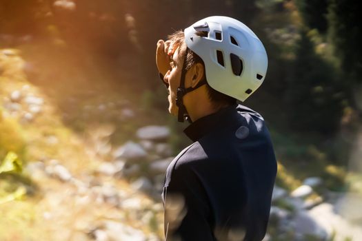 A young man in a white mountaineering helmet looks at the trail along the route of climbing the mountains in the rays of the sun. A traveler makes an ascent to a mountain in a national park.