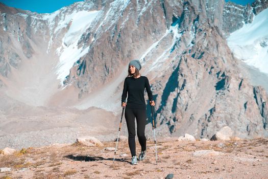 A young beautiful sports girl in thermal underwear with trekking poles walks along the trail among the snow-capped high peaks of the mountains, the traveler walks and climbs tin the national park.