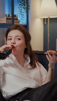 Person eating slice of pizza while watching comedy on TV and laughing on couch at home. Caucasian woman having fun with movie on television, enjoying fast food takeaway and bottle of beer