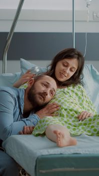 Caucasian man and woman expecting child in hospital ward while african american nurse fixing IV drip bag for pregnant mother at maternity. Young happy couple waiting on giving birth