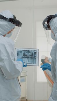 Stomatologists with ppe suits holding digital tablet with teeth x ray analyzing dental healthcare for implant operation at medical clinic. Dentists using scan technology during pandemic