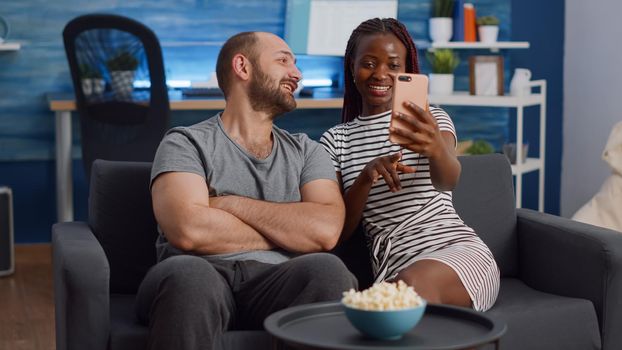 Modern interracial couple talking on video call conference at home. Young multi ethnic partners using internet for remote communication, sitting together on couch in living room.