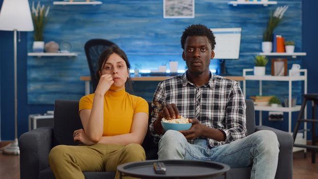 POV of interracial couple watching movie on TV with snack at home. Married multi ethnic lovers looking at camera while eating popcorn in living room. Mixed race people bonding on sofa
