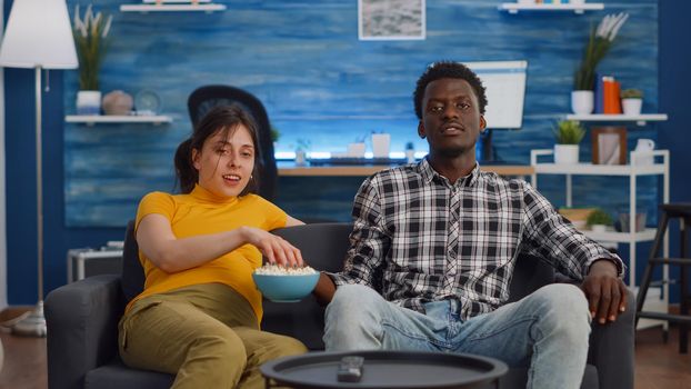 POV of interracial couple with pregnancy watching TV and eating snack on couch in living room. Young multi ethnic partners expecting baby and sitting on sofa at home with popcorn