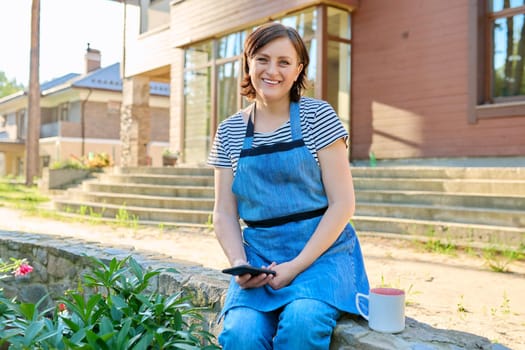 Portrait of happy middle aged woman relaxing outdoors in backyard. Female in garden apron with mug of tea and smartphone in her hands, looking at camera, spring garden background. 40s people lifestyle