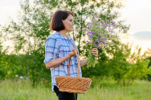 Middle-aged smiling woman with basket and bouquet of wildflowers in nature. Rural outdoor portrait of 40s female, in spring summer meadow. Beauty, people, nature, women concept