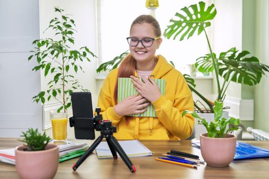 Teenage girl looking at webcam using smartphone for an online lesson, reading textbook book. Student 12, 13 years old, studying remotely at home, e-learning, virtual lessons, distance learning