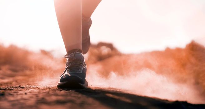 Female athletic legs in sneakers at sunset during outdoor jogging at sunset. A young girl warms up, she is engaged in trail running, view on her legs and clouds of dust close-up.