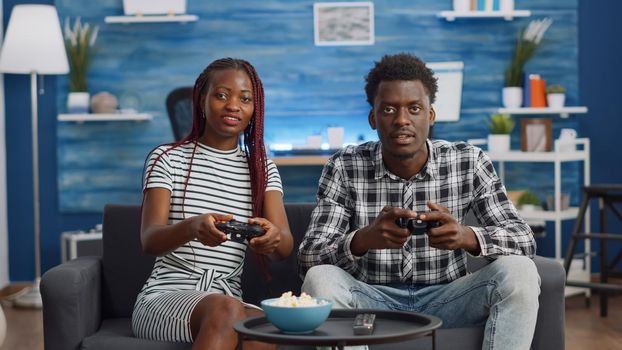 African american people playing video game with joysticks at home on sofa. Black couple enjoying fun activity with console and television, sitting on living room couch. Cheerful relationship