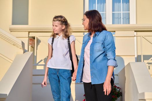 Mother and daughter schoolgirl with backpack on porch of house. Mom holding preteen student by hand as she goes to school on sunny morning. Parent child relationship, family, back to school concept