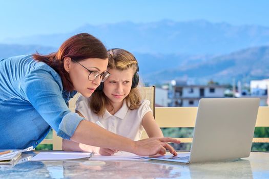 Child girl distance learning online with laptop, mom helps. Preteen in headphones with notebooks, talking to mother, sitting on home outdoor terrace. E-education, virtual lessons, technology school