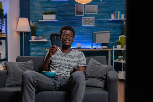 African american guy resting on couch watching comedy film on television laughing during movie enjoying free time in living room. Young man sitting on life relaxing while eating popcorn