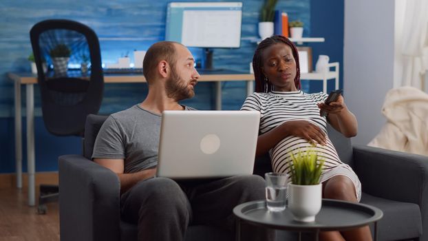 Young interracial partners with pregnancy using technology at home. Caucasian man using digital laptop while african american pregnant wife watching television sitting in living room