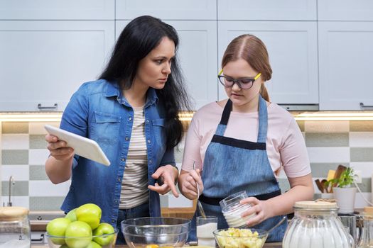 Mom and teen daughter preparing apple pie together, at home in kitchen. Woman with digital tablet, recipe, video call, technology in daily life. Family, parent teenager relationship, lifestyle