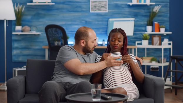 Interracial couple with pregnancy watching movie on TV while eating popcorn. Multi ethnic parents expecting baby and sitting on living room couch. Mixed race people relaxing with television