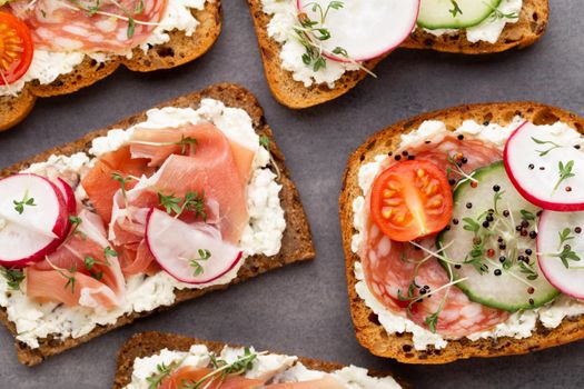 Variety of mini sandwiches with cream cheese, vegetables and salami. Sandwiches with cucumber, radish, tomatoes, salami on a gray background, top view. Flat lay.