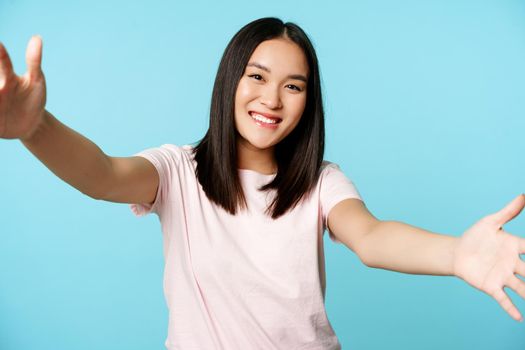 Portrait of smiling asian woman reaching for hug, spread hands friendly, taking, holding smth large, standing over blue background.