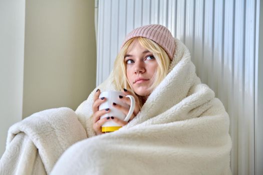 Heating during the cold winter autumn season. Young female teenager woman warming herself with a blanket hat, a central heating radiator, hot tea