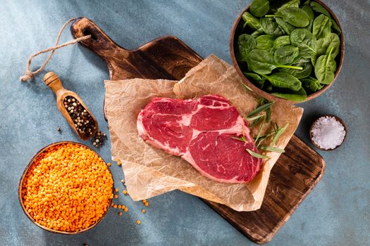 Fresh raw rib-eye steak on wooden cutting board, with spinach, lentils and rosmary in a rustic style.