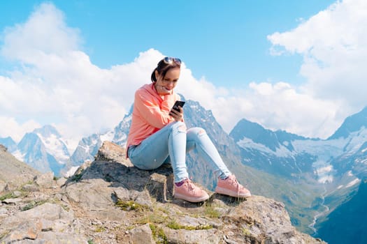 Full body woman sitting on rock and browsing smartphone against cloudy sky on sunny day in mountains