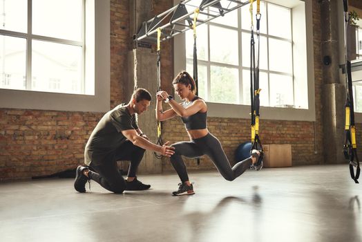 Doing squat exercise. Confident young personal trainer is showing slim athletic woman how to do squats with Trx fitness straps while training at gym. TRX Training. Exercising together. Active lifestyle