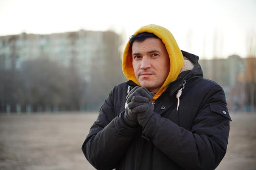 Young frozen man in yellow hood and black jacket. Adult male warming hands in gloves with his breath in cold weather