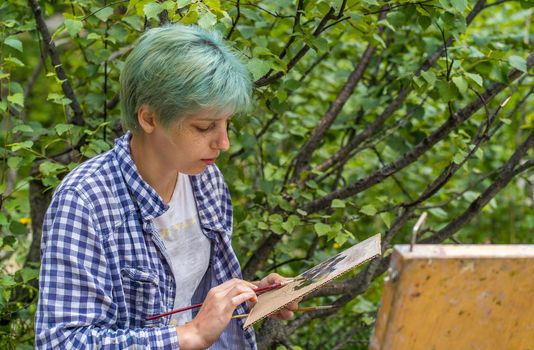 Close up of young woman mixes paints in palette, drawing picture on cardboard on hike. Paintress mixes colors, drawing mountain landscape.