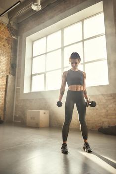 Working with weight. Young athletic woman in sportswear exercising with dumbbells while standing in front of window at gym. Professional sport. Workout. Healthy lifestyle