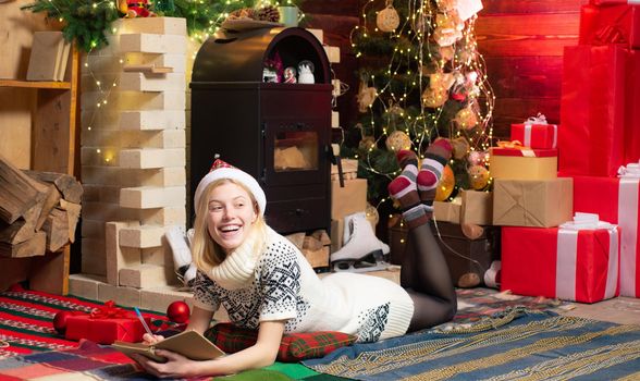 Teenage blonde girl in knitted sweater dress and red hat writing a wish list of presents to Santa. Christmas magic concept. Happy winter holidays at cozy decorated home with family