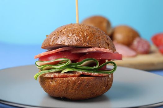 burger sandwich of buckwheat dark bun sausage salami vegetables cucumbers tomato on a gray plate on a blue background. Homemade instant food.