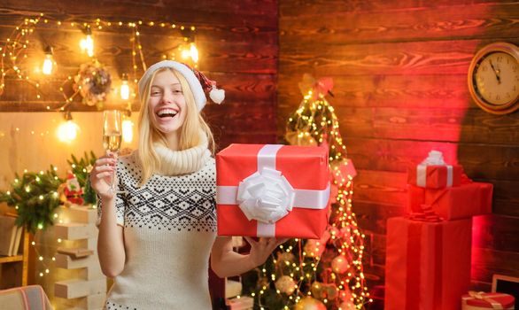 Happy blonde woman in Santa's red hat holding a present gift in one hande and a widow champagne in another at beautiful decorated interior. Celebrating of New Year concept