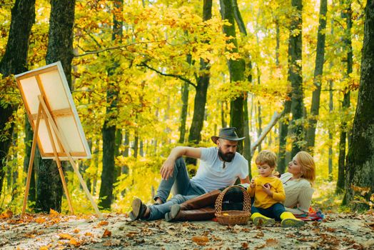 Young family of artists doing a picnic on an autumns day. The concept of a happy family. Young smiling family doing a picnic on an autumns day. Drawing easel with artwork. Inspiration