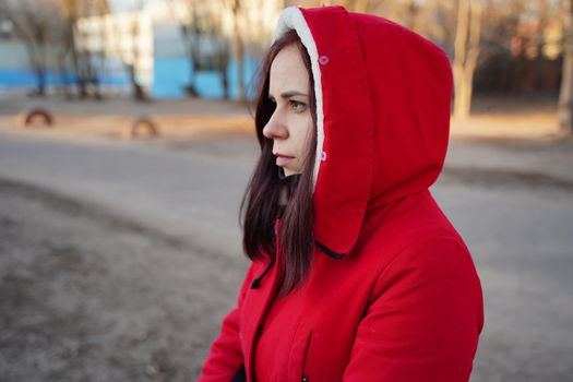 Portrait of young woman in red hood and jacket on street. Adult serious female looks away thoughtfully, standing on fresh air