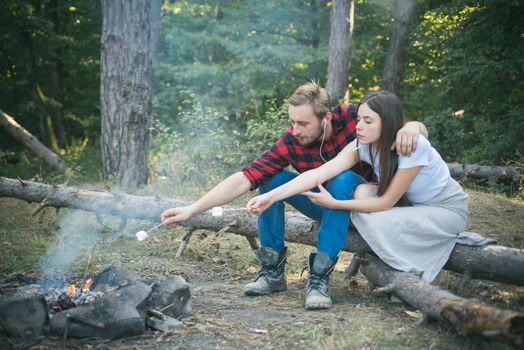 Pretty couple roast marshmallow candies on the campfire in forest. Tourists relaxing. Picnic time. Tourism concept. Young couple having picnic in woods. Camp fire in summer