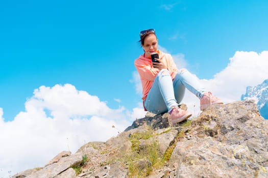 Full body woman sitting on rock and browsing smartphone against cloudy sky on sunny day in mountains