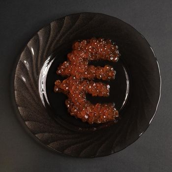 The symbol of the euro is beautifully assembled with real red caviar on a black plate on a gray background view from above. Food in the world's elite restaurants is a symbol of money