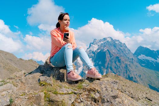 Full body woman sitting on rock and browsing smartphone against cloudy sky on sunny day in mountains