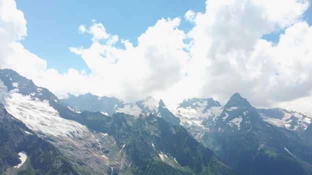 From below white clouds floating on blue sky over mountain ridge covered with snow on sunny day in nature