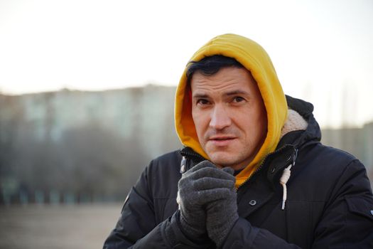 Young frozen man in yellow hood and black jacket. Adult male warming hands in gloves with his breath in cold weather