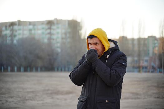 Young frozen man in yellow hood and black jacket. Adult male warming hands in gloves with his breath in cold weather