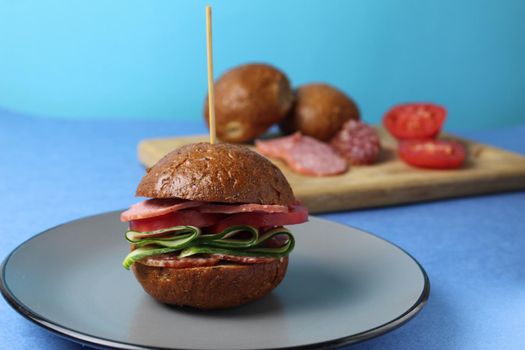 burger sandwich of buckwheat dark bun sausage salami vegetables cucumbers tomato on a gray plate on a blue background. Homemade instant food.