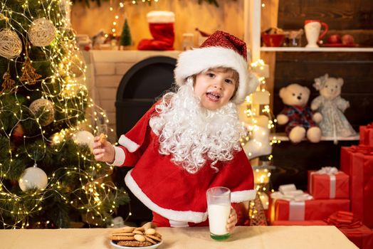 Happy new year. Santa in home. Little Santa Claus kid with beard and mustache. Cheerful Santa Claus holding glass with milk and cookie with fireplace and Christmas Tree in the background